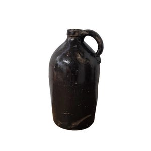 May include: A dark brown ceramic jug with a handle. The jug has a slightly textured surface and a rounded body.