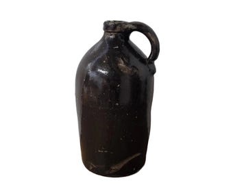 May include: A dark brown ceramic jug with a handle. The jug has a slightly textured surface and a rounded body.