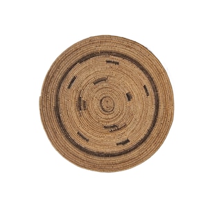 May include: A round, woven basket with a natural brown color and a darker brown circular design. The basket is made of tightly woven fibers, creating a textured surface. The design features a series of rectangular shapes around the center.