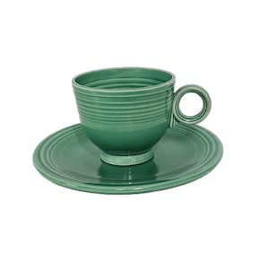 May include: A sage green ceramic teacup and matching saucer set. The teacup features a rounded handle and horizontal ribbed detailing around the exterior. The saucer has a circular design with concentric rings embossed along the outer edge.