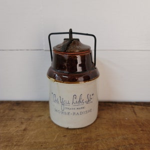 May include: A brown and white ceramic jar with a metal wire handle. The jar has a label that reads "As You Like It (Trade Mark) Horse-Radish."