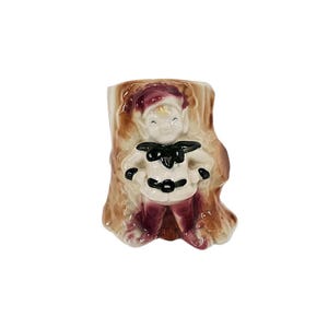 May include: A ceramic wall planter shaped like a tree trunk with a small elf figure standing inside. The elf is wearing a red hat, a white shirt with a black belt, and brown pants. The planter is painted in shades of brown, red, and white.