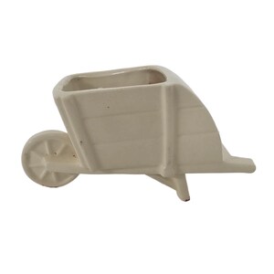 May include: A cream ceramic wheelbarrow planter. The planter features a rectangular trough and a single wheel. The wheelbarrow is designed with horizontal lines and a small handle. It is a decorative item for small plants.