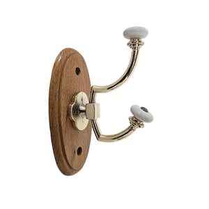 May include: A wall-mounted coat hook featuring an oval-shaped light brown wooden backplate with two screw holes. The hook is made of polished gold-tone metal with two curved arms, each topped with a small, smooth, white ceramic knob at the end.