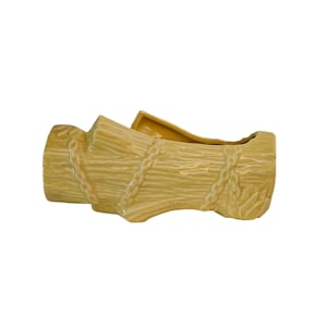 May include: A ceramic planter shaped like a bundle of wheat stalks, tied with rope. The planter is a light yellow colour with a textured surface, and is designed to hold plants or flowers.