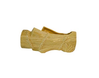 May include: A ceramic planter shaped like a bundle of wheat stalks, tied with rope. The planter is a light yellow color with a textured surface, and is designed to hold plants or flowers.