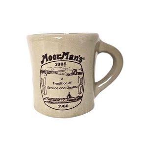 May include: A beige ceramic coffee mug with a brown logo that reads "MoorMan's 1885 A Tradition of Service and Quality 1980".
