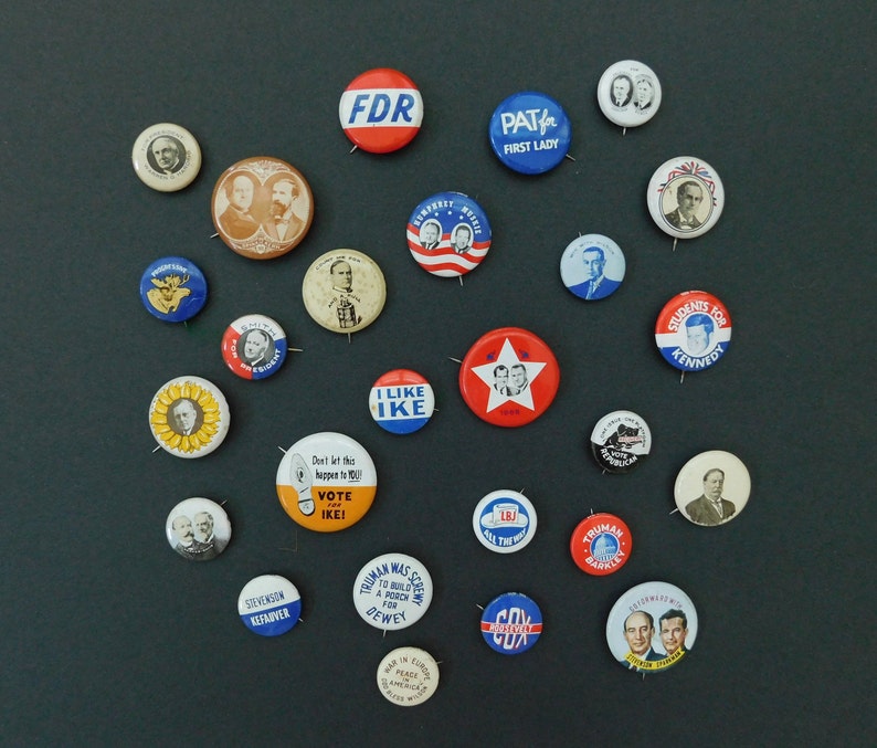 May include: A collection of vintage political campaign buttons featuring various candidates and slogans. The buttons are circular and made of metal with a variety of colors and designs. Some of the buttons feature portraits of candidates, while others feature slogans or logos. The buttons are arranged in a circular pattern on a gray background.