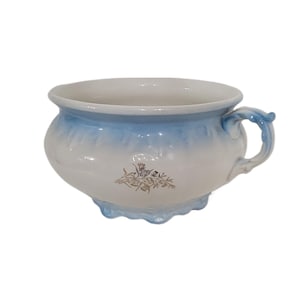 May include: A white and blue ceramic chamber pot with a handle. The pot has a rounded shape with a blue ombre effect around the rim and base. A floral design is featured on the front.
