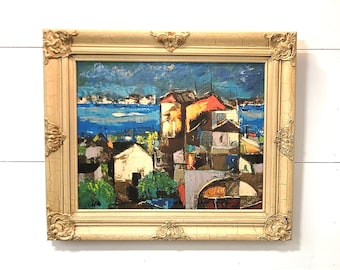 May include: A framed abstract painting depicting a cityscape with colorful buildings and a blue body of water in the background.