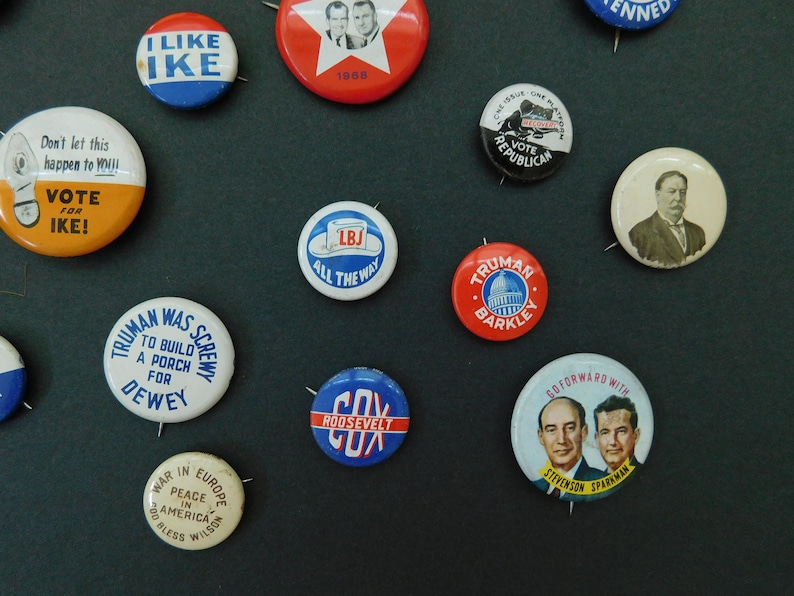 May include: A collection of vintage political campaign buttons featuring various candidates and slogans. The buttons are round and made of metal with a pin on the back. Some of the buttons feature images of candidates, while others feature text slogans. The buttons are arranged in a random pattern on a dark background.