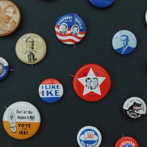 May include: A collection of vintage political campaign buttons featuring various candidates and slogans. The buttons are mostly round and feature red, white, and blue colors. Some buttons have images of candidates, while others have text slogans. The buttons are arranged on a dark surface.