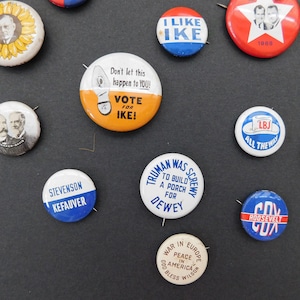 May include: A collection of vintage political campaign buttons featuring various candidates and slogans. The buttons are round and made of metal with colorful designs and text. Some of the slogans include "Vote for Ike", "I Like Ike", "Truman Was Screwy", "LBJ All The Way", and "Peace in America".