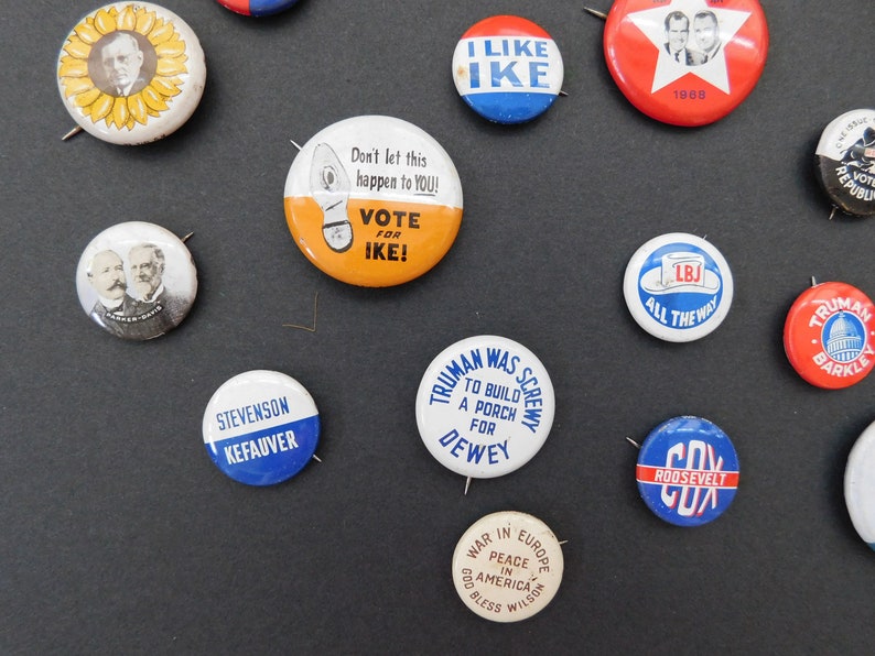 May include: A collection of vintage political campaign buttons featuring various candidates and slogans. The buttons are round and made of metal with colorful designs and text. Some of the slogans include "Vote for Ike", "I Like Ike", "Truman Was Screwy", "LBJ All The Way", and "Peace in America".