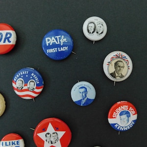 May include: A collection of vintage political campaign buttons featuring various candidates and slogans, including "FDR", "PAT for FIRST LADY", "HUMPHREY MUSKIE", "WIN WITH WILSON", and "STUDENTS FOR KENNEDY".