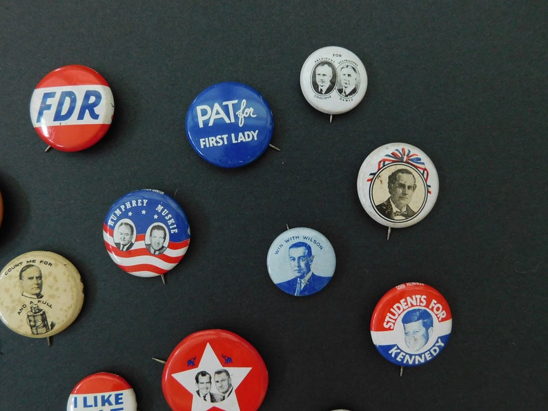 May include: A collection of vintage political campaign buttons featuring various candidates and slogans, including "FDR", "PAT for FIRST LADY", "HUMPHREY MUSKIE", "WIN WITH WILSON", and "STUDENTS FOR KENNEDY".