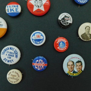 May include: A collection of vintage political campaign buttons featuring various candidates and slogans. The buttons are round and made of metal with colorful designs and text. Some of the buttons feature portraits of candidates, while others have text-only slogans. The buttons are arranged on a dark background.