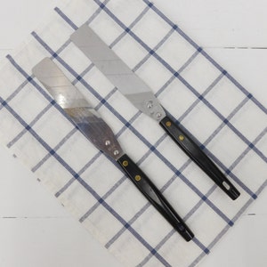 May include: Two black handled metal spatulas with silver blades on a white and blue checkered surface.