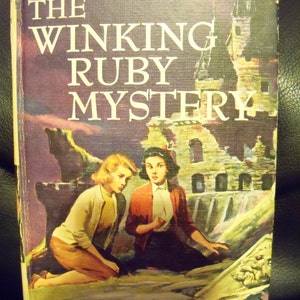May include: A vintage paperback book cover with a purple background featuring the title "The Winking Ruby Mystery" in white text. The cover depicts two young women standing in front of a crumbling stone building with a large window. The book is part of "The Dana Girls Mystery Stories" series and is written by Carolyn Keene.