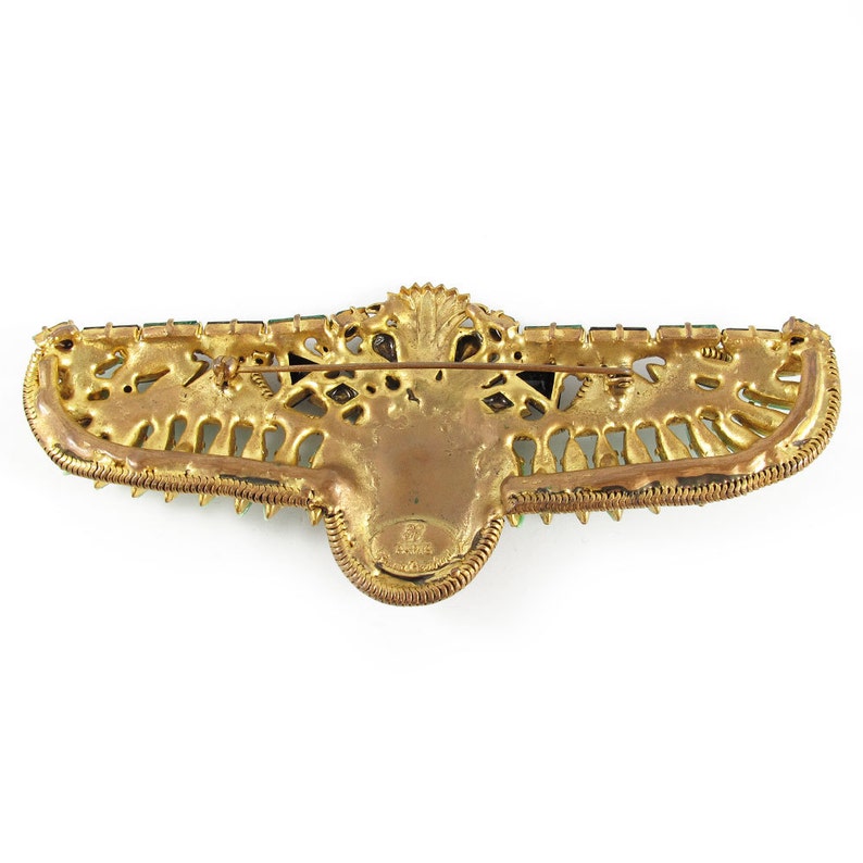May include: Gold-tone brooch with green and black rhinestones, shaped like a winged creature with a crown on its head. The back of the brooch has a signature that reads "Schiaparelli".