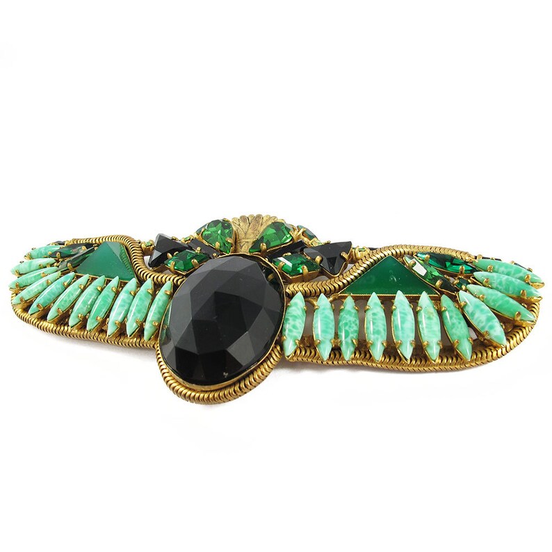 May include: A gold-tone brooch with a large black faceted stone surrounded by green and clear faceted stones. The brooch is shaped like a winged creature.