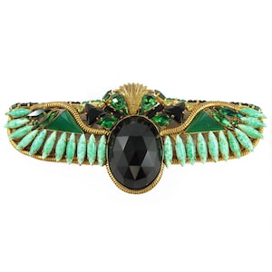May include: A gold-toned brooch with a large black faceted stone surrounded by green and black rhinestones. The brooch is shaped like a winged scarab beetle.