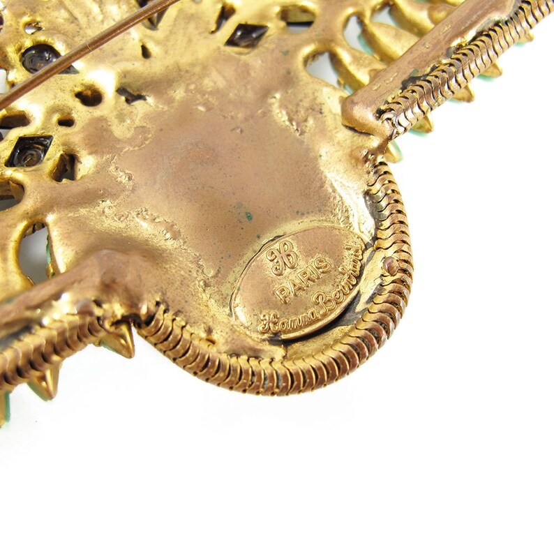 May include: Gold-toned metal brooch with a detailed design and green rhinestones. The back of the brooch has a circular label that reads "96 PARIS 96" in a gold font.