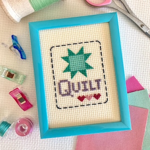 May include: A framed cross-stitch design with a green star and the word "QUILT" in purple letters. There are three pink hearts below the word. The frame is a bright turquoise blue.