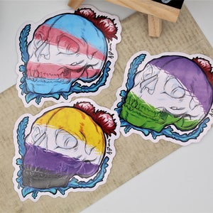 Puede incluir: Tres pegatinas con diseños de calaveras con esquemas de colores de banderas del orgullo. Las calaveras tienen esquemas de colores azul, rosa y blanco, morado, verde y blanco, y amarillo, blanco y morado. Cada calavera está decorada con una flor roja y hojas azules.