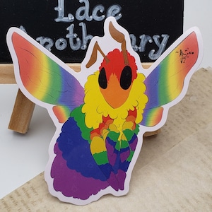Puede incluir: Una pegatina de polilla de color arcoíris con un cuerpo amarillo peludo y una cabeza roja. La polilla tiene alas grandes con un degradado arcoíris y un contorno negro. La pegatina está sobre un fondo blanco.