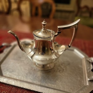 May include: A silver teapot with a fluted design sits on a silver tray with a hammered finish. The teapot has a lid and a curved handle.