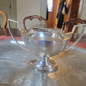 May include: A silver sugar bowl with a decorative design and two handles. The bowl is sitting on a silver tray with an embossed floral design.