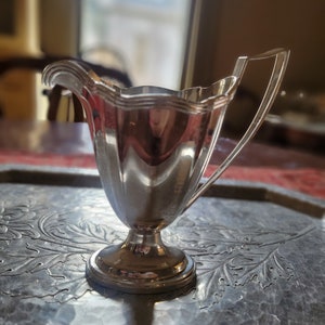 May include: A silver creamer with a fluted design and a handle. The creamer is sitting on a silver tray with an embossed floral pattern.