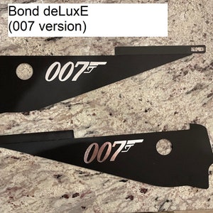 Könnte beinhalten: Zwei schwarze Metallplatten mit dem Text "007" in silberner Schrift. Die obere Platte hat den Text in einer größeren Schriftgröße und die untere Platte hat den Text in einer kleineren Schriftgröße. Der Text "Bond deLuxE (007 Version)" ist in weißer Schrift auf weißem Hintergrund geschrieben.