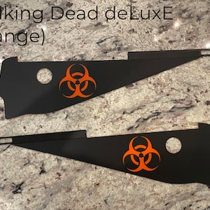 May include: Two black metal side panels for a pinball machine with an orange biohazard symbol. The text "Walking Dead deLuxE (orange)" is visible at the top of the image.