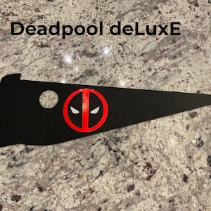 May include: Black and red Deadpool pennant with a white eye mask. The pennant is shaped like a triangle with a rounded top and a pointed bottom. The text "Deadpool deLuxE" is printed in white at the top of the pennant.