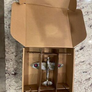 May include: A cardboard box with a model airplane inside. The airplane is a green, brown, and white camouflage color scheme with red and white markings. The box has a divider to keep the airplane in place.