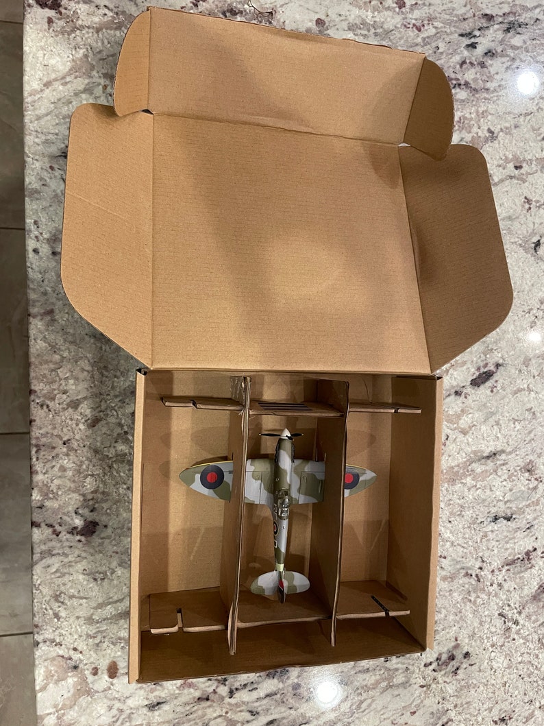 May include: A cardboard box with a model airplane inside. The airplane is a green, brown, and white camouflage color scheme with red and white markings. The box has a divider to keep the airplane in place.