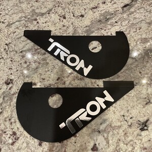May include: Two black metal pieces with a silver "TRON" logo. The pieces are shaped like a crescent moon and have a hole in the center.