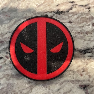 Może przedstawiać: Okrągłe logo w kolorze czerwonym i czarnym ze stylizowanym wzorem oka. Logo przedstawia popularną postać z komiksów Marvela.