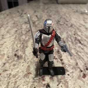 以下が含まれることがあります： 銀色のヘルメットと鎧、赤いバンドリエール、黒いズボンを身に着けたマンダロリアンアクションフィギュア。フィギュアは銀色の剣とブラスターを持っています。黒い台座の上に立っています。