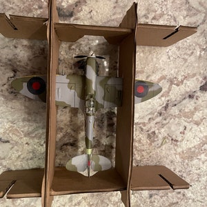 May include: A model of a green, gray, and brown camouflage World War II era fighter plane with red and white markings. The plane is in a cardboard box.