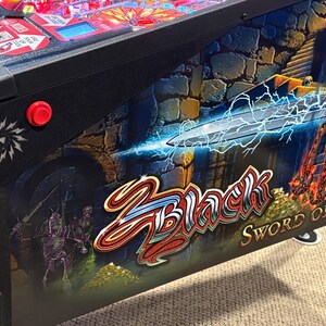 Op de afbeelding: Een flipperkast met een zwarte kast en een kleurrijk speelveld met een zwaard, bliksem en de tekst "Black Knight Sword of Rage".