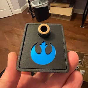 May include: A black plastic wall mount with a blue Rebel Alliance symbol and a wooden dowel. "MADE IN CHINA" is printed on the box in the background.