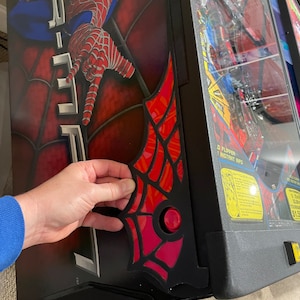 May include: A black and red Spider-Man pinball machine with a red web design on the side. The machine features a red button and a clear plastic cover. The Spider-Man logo is visible on the side.