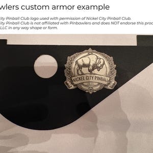 May include: A black, flat, custom-shaped piece of pinball armor featuring a circular cutout and a metallic emblem. The emblem displays a buffalo illustration surrounded by a wreath and the text Nickel City Pinball 2025 in a circular frame.