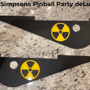 May include: Two black metal pinball machine side rails with yellow radiation symbol decals. The Simpsons Pinball Party deLuxE is written in white text at the top of the image.