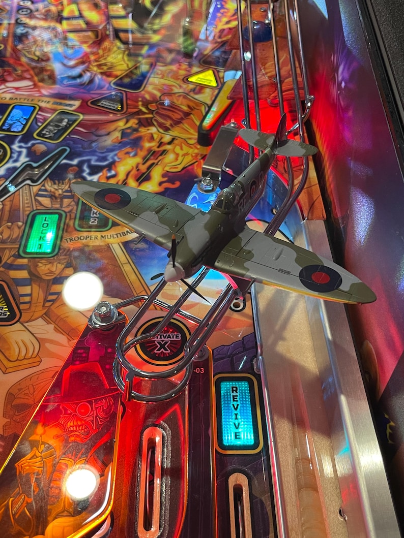 May include: A green and brown toy airplane with red and white markings is on a pinball machine. The airplane is on a metal ramp and is facing the right side of the image. The pinball machine has a red and orange background with a yellow and black warning sign. The pinball machine also has a blue and yellow "Revive" button.