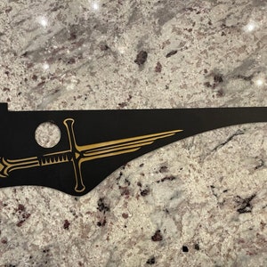 May include: A black and gold sword design on a black background. The sword is stylized and has a long, curved blade. The design is on a piece of wood or plastic.