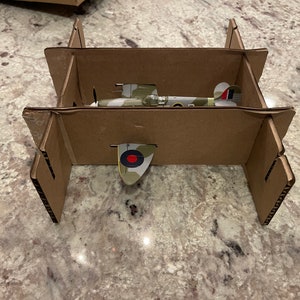 May include: A cardboard box with a model airplane inside. The airplane is painted in camouflage colors with a red and white roundel on the tail.